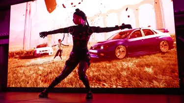 A performer on stage in front of a screen image of two cars in a field and a figure mimicking the movement of the performer