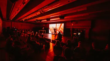 An audience watching a performer in front of a screen image under a dark red light
