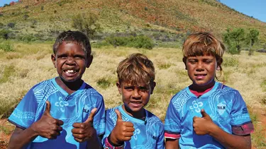 Three children wearing blue jerseys give thumbs up, smiling broadly in a grassy field with a mountain in the background under a clear blue sky.