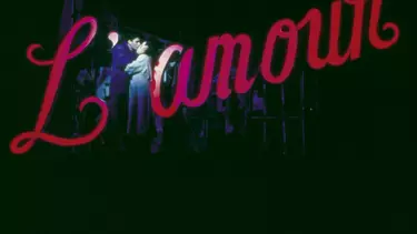 A couple embraced in a kiss, against a black background, standing in front of red lit life-size lettering that reads 'L'amour'.