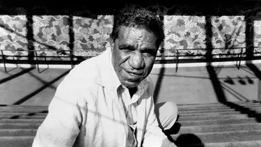 Jagamara photographed in black and white sitting on carpeted steps in front of the mural 'Possum Dreaming'.