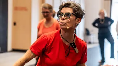 A woman training a seniors class through movement. She is wearing a red top and a microphone headset.