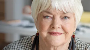 Elderly woman with white hair and plaid outfit smiles warmly at the camera.