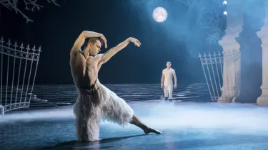 A topless man in a white feathery costume dancing under a set with a full moon on a blue lit stage.