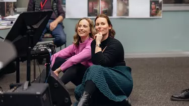 Two women sit on the floor in a rehearsal room, they are smiling and are seated next to a keyboard.