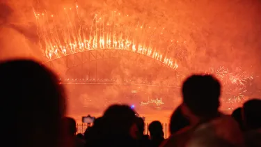Point of view crowd watching yellow and orange smokey fireworks over the Sydney Harbour Bridge.