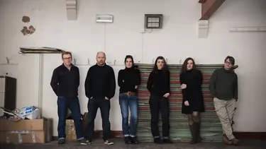 Six people wearing dark colours standing against a wall.