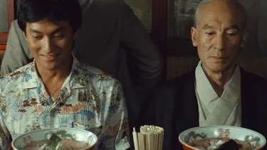 Two men are seated side by side at a restaurant with bowls of food in front of them. One man is younger and dressed casually, the other is older and dressed formally.