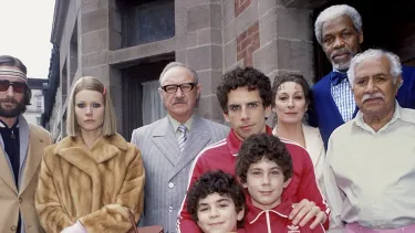 Cast members from The Royal Tenenbaums stand together outside a large house. They are all posing in a formal group portrait.