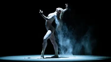 A dancer is illuminated by a spotlight in a dark room. She is covered in a white dust that is falling off her shoulders and hair. Her arms are outstretched.