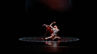 Two dancers wearing limited clothing are sitting on the ground in the middle of a spotlighted circle.