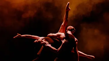 A dancer in a black singlet is facing away holding up another dancer who is outstretched in an acrobatic pose. The room is hazy and illuminated in an orange hue.