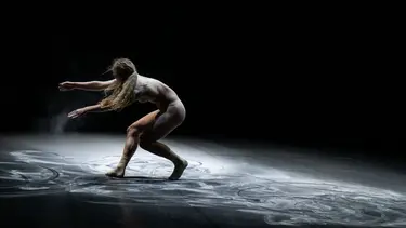 A dancer moves across a dusty floor. They are bent over with their arms stretched outwards.