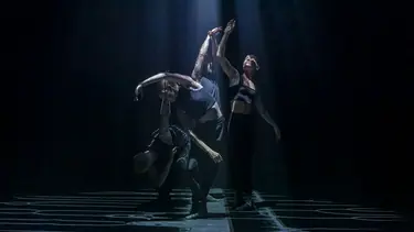 A group of four dancers wearing black standi in close proximity in various poses. Streams of light illuminate them from above.