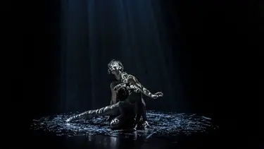 Two dancers are on a black floor illuminated by streams of light coming from above.