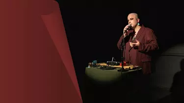 A bald person in a coat speaking into a mic.