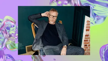 A white man in a casual grey suit and glasses sits relaxed in a chair. The background is an iridescent purple swirl, looking like water.