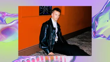 A man sits on a carpeted floor against an orange wall. He is wearing a black leather jacket and black trousers. The background is a purple, iridescent swirl, looking like water.