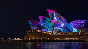 The Sydney Opera House sails are visible with the harbour in the foreground. The sails are illuminated with projected art featuring a variety of colourful elements.