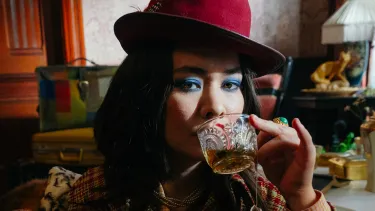 A woman in a red hat and plaid coat with bright blue eyeshadow drinks from a glass, surrounded by eclectic, vintage decor and a retro telephone.
