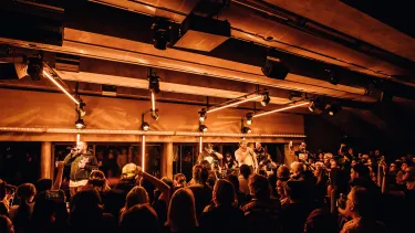 Four men are performing on a stage under lights. A crowd is looking on.