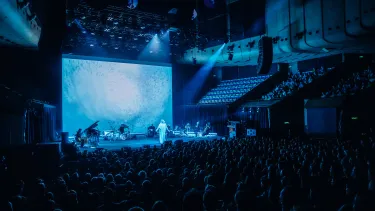A performer is centre stage backed by a band and string section. There is a large screen projecting an image behind them. A seated crowd watches on.