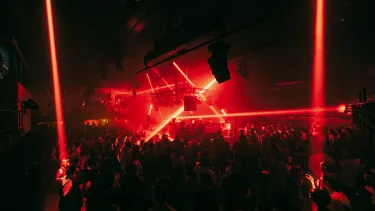 Red light beams shine sporadically across a crowded room that has a raised dj stage at its center.