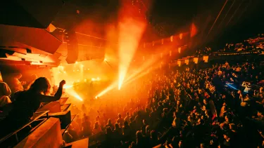 A tiered crowd watch a stage occupied by a band with lights shooting out in different directions. One crowd member is leaning over a balcony for a better view.