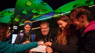 A group of students touch a Sydney Opera House tactile model in front of the sails lit up with a green pattern.