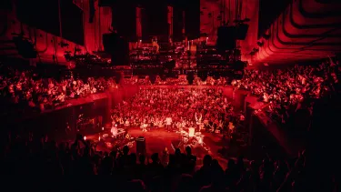 A band is seen from above. They are lit up in a red hue surrounded by a standing and applauding crowd from all angles.