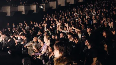 A crowd is standing, some are dancing, some have their arms in the air and others are filming with their phones.