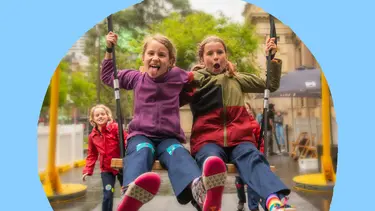 Two young girls sit side by side on a swing. Their legs are outstretched and they have smiling faces.