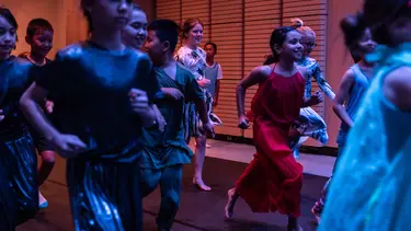A group of children are running along a black floor with a wooden wall behind them.