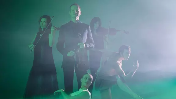A white, 50-year old man holds a violin immersed in green light. Around him is two female violinists, a man dancer and a female ballerina.