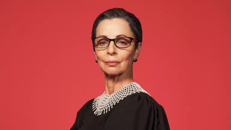 A white woman in her 60s has her hair sleekly pulled back and pursed lips wears black glasses and a lawyer's black robe with white crocheted neck piece. She stands in front of an all-red background.