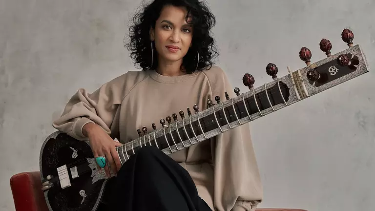 An Indian woman with short, curly black hair wears big silver hoop earrings and a beige long sleeve top. She has a sitar placed across her knee - this is a guitar-like instrument with a short round body and a very long neck and many tuning knobs.