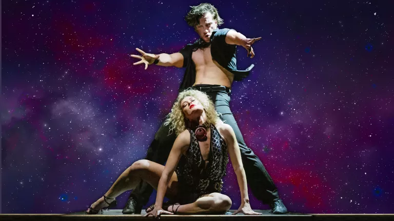 A male dancer wearing black and an open button shirt stands behind a female dancer sitting on the floor.