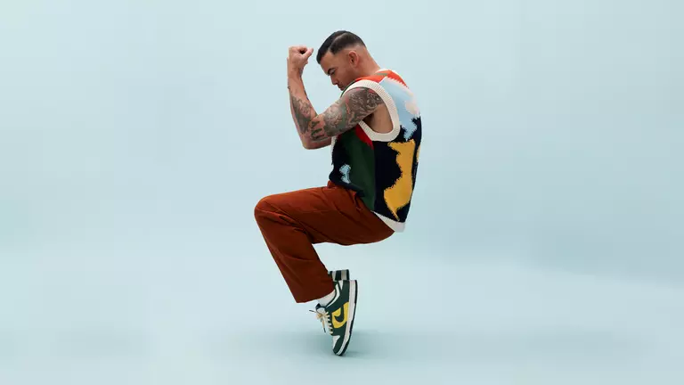 A man with tanned skin and short black hair is posed standing on his toes, with bent knees and his arms held up in front of his face. He wears maroon pants, green and yellow Nike shows and a multi coloured vest.