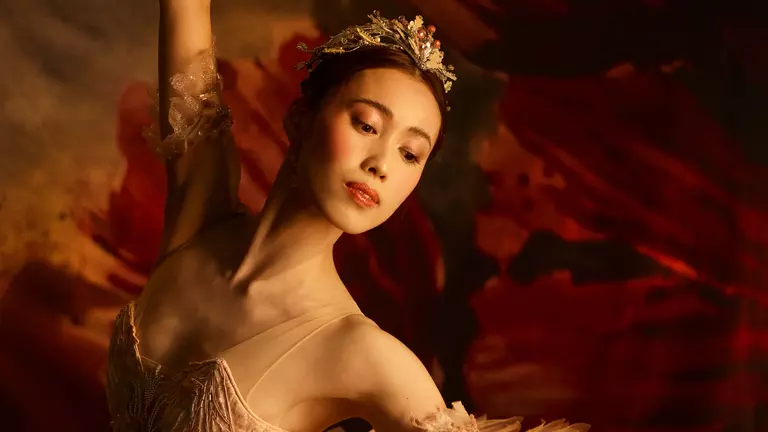 A ballerina with brown hair wears a gold tutu and tiara with gold arm cuffs. She looks over her shoulder in front of a red background.