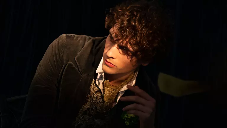 A man with light brown curly hair looks off into the distance in front of a black background. He wears a gold tie, white shirt and green jacket.