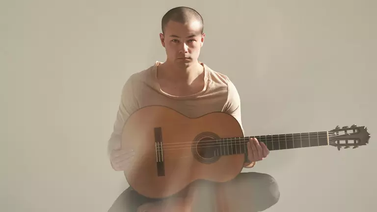 A young Asian man with shaved hair wears a beige top and holds an acoustic guitar.