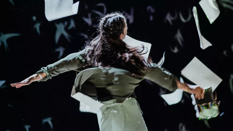 A women dances surrounded by falling paper. She is holding a book and her hair is in braids.