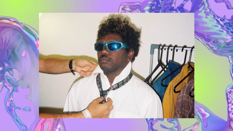 An Indigenous man in sunglasses is casually helped with a tie. The background is a purple, iridescent swirl, looking like water.