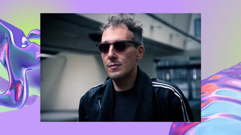 A white man wears sunglasses and a dark jacket. Background of a purple, iridescent swirl, looking like water.