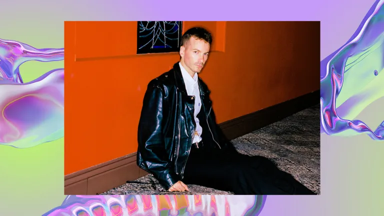 A man sits on a carpeted floor against an orange wall. He is wearing a black leather jacket and black trousers. The background is a purple, iridescent swirl, looking like water.