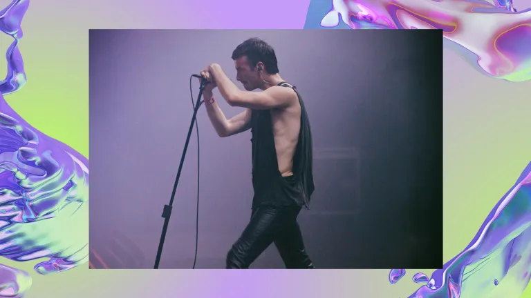A male singer passionately grips a microphone on a stand during a performance. He wears skinny trousers and a flowy tank top. The background is a purple, iridescent swirl, looking like water.