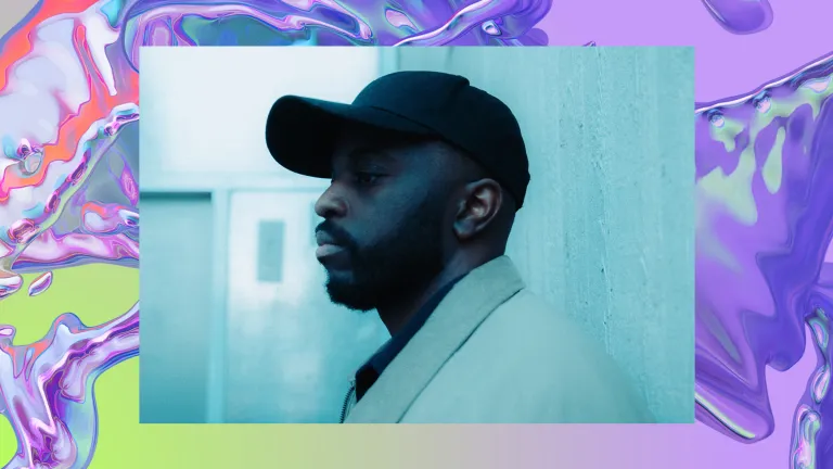 Alfa Mist stands against a concrete wall wearing a black cap.