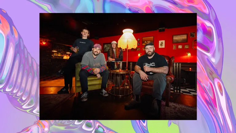 Four men sit on cozy, vintage couches in a cozy, dimly lit room with red walls and warm lighting. The image overlays a background of a purple iridescent swirl, looking like water.