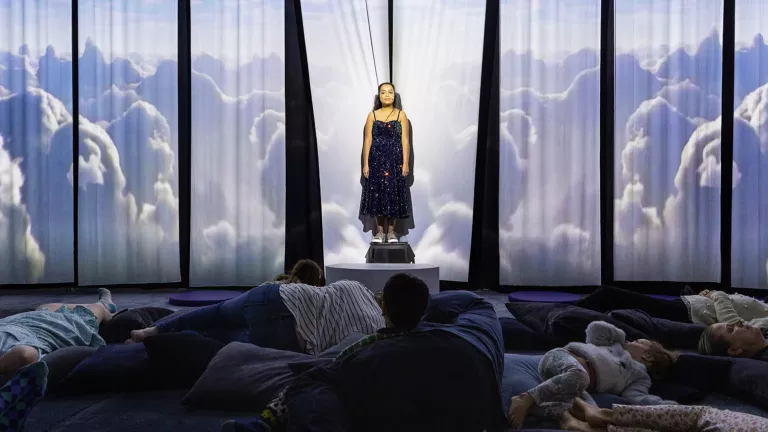 A young white woman with dark brown hair stands in front of a fabric background, where projections of clouds are being cast. An audience of meditation children and adults lie on pillows in front of her.