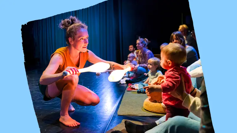 A woman holds two glowing juggling pins while a child sits on the floor, both engaged in a playful moment together.